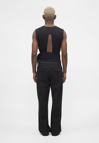 Άτομο με κοντό ξανθό μαλλί, που φοράει μαύρο αμάνικο τοπ με άνοιγμα στην πλάτη και μαύρο παντελόνι, στέκεται με την πλάτη προς ένα απλό φόντο.