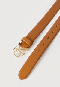 Καφέ δερμάτινη ζώνη με χρυσή αγκράφα "CK" και πολλαπλές τρύπες ρύθμισης, τοποθετημένη επίπεδη σε λευκή επιφάνεια.