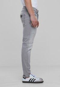 Szürke, slim-fit farmernadrág, koptatott részletekkel, oldalsó zsebekkel és fekete logólogóval. Fehér sportcipővel viselve, fekete csíkokkal.