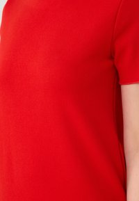 Gros plan sur le bras d'une femme portant une manche d'une chemise rouge vif, mettant en valeur la texture et la couleur du tissu.