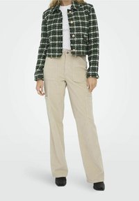 Femme portant un pantalon beige à jambes larges, un haut blanc et une veste courte à carreaux verts et blancs avec des boutons, debout devant un fond uni.