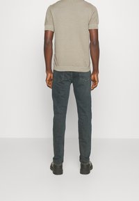 Camisa de punto de manga corta texturizada en color beige con cuello redondo, combinada con jeans azul oscuro y botas negras, vista desde atrás.