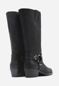 Botas de cuero negro hasta la rodilla con punta redonda, cremallera lateral y una correa con hebilla adornada con tachuelas metálicas. Tacón bajo y apilado.