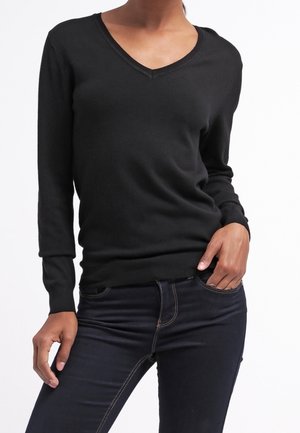 Kobieta ubrana w dopasowany czarny sweter z długim rękawem i dekoltem w serek oraz ciemnoniebieskie dżinsy, z jedną ręką w kieszeni, na białym tle.