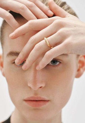 Anillo de oro con una superficie lisa y reflectante, llevado en una mano; los dedos son delicados y están parcialmente ocultos por otra mano situada encima.
