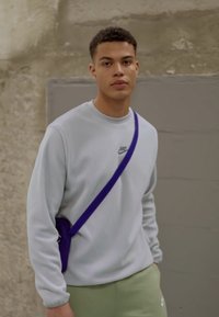 Lys grå hettegenser med en liten Nike-logo, kombinert med lysegrønne bukser. En lilla skulderveske bæres over kroppen. Nøytral bakgrunn.