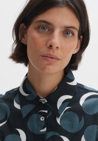 Camicia con colletto in tessuto blu scuro caratterizzata da un motivo astratto ripetuto in azzurro chiaro e bianco. Chiusure a bottoni sul colletto.