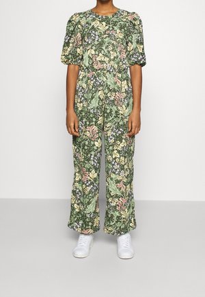 Personne portant une combinaison verte à motif floral avec des manches bouffantes et des baskets blanches, debout devant un fond uni.