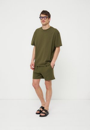 Homme portant un t-shirt oversize vert olive et un short avec des sandales noires à boucle, debout devant un fond blanc uni.