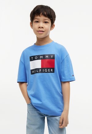 Ragazzo in piedi con una mano in tasca, indossa una maglietta blu Tommy Hilfiger e jeans azzurri, su uno sfondo bianco uniforme.