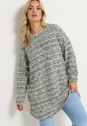 Pitkähiuksinen nainen, jolla on kultaiset renkaankorvakorut, harmaa-valkoinen teksturoidusta neuloksesta valmistettu neulepaita ja vaaleansiniset farkut, poseeraa valkoista seinää vasten.