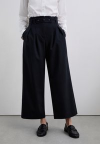Black pinstriped pleated trousers with a high waistband, featuring three buttons and side pockets, paired with black loafers.