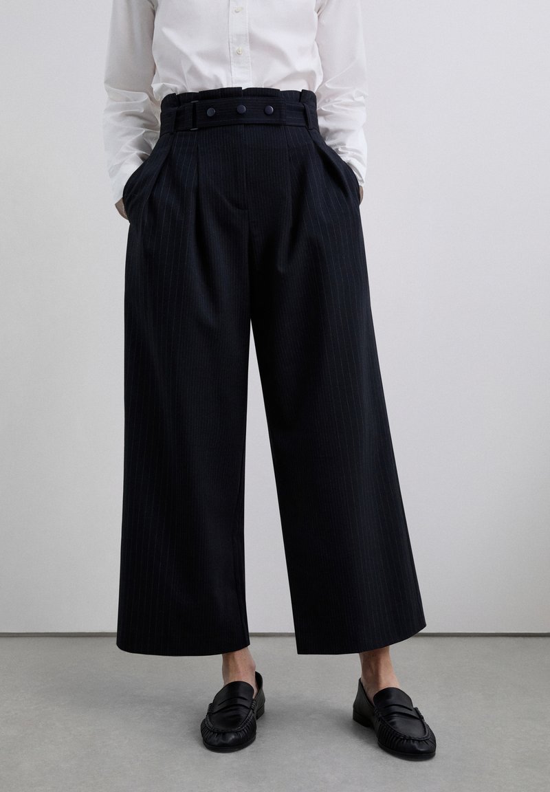 Black pinstriped pleated trousers with a high waistband, featuring three buttons and side pockets, paired with black loafers.
