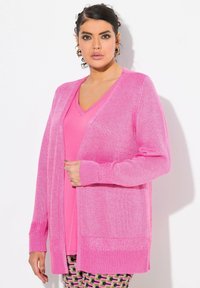 Cardigan léger rose avec un design en tricot texturé et des poignets côtelés ; porté ouvert sur un top rose assorti et un pantalon à motifs.