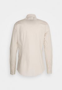 Beige långärmad skjorta med knappar, sedd från baksidan, med klassisk krage och knäppta manschetter mot en enfärgad vit bakgrund.