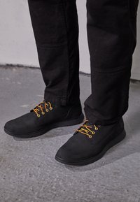 Zapatos negros con un acabado liso, cordones amarillos y una suela texturizada, combinados con pantalones negros, sobre un suelo gris.