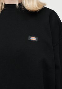 Ξανθό άτομο που φοράει μαύρο φούτερ με στρογγυλή λαιμόκοψη και ένα μικρό ορθογώνιο λογότυπο Dickies στο αριστερό στήθος.