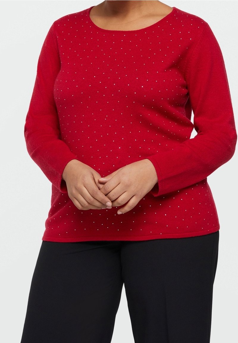 Femme portant un pull rouge à manches longues avec de petits points blancs, debout, les mains jointes, portant un pantalon noir.
