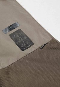 Innenansicht einer wasserdichten Performance-Jacke, ausgestattet mit hellbraunem Stoff, strukturiertem Muster sowie Pflegeetiketten mit schwarzem Text und Reißverschluss-Details.