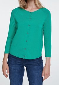 Grüne Strickjacke mit Dreiviertelärmeln, einem runden Ausschnitt und einer Knopfleiste. Auffällige graue Knöpfe auf der Vorderseite.