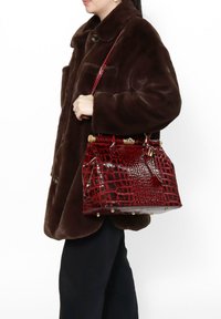 Borsa a mano rossa con motivo coccodrillo e finitura lucida, dettagli in hardware dorato e design strutturato, tenuta da una persona in un cappotto marrone.