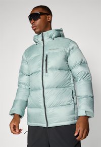 Light green puffy jacket with a zippered front, adjustable hood, and side pockets. Features a chest pocket and Columbia logo embroidered.