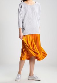 Grå långärmad topp med vid halsringning, matchad med en ljusorange plisserad kjol och vita sneakers. Kjolen har ett flödande, strukturerat utseende.