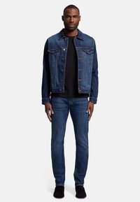 Jeansowa kurtka w ciemnym niebieskim kolorze noszona na czarnej koszuli, w zestawieniu ze średnio niebieskimi dżinsami. Klasyczny fason z dwiema kieszeniami na piersi, metalowymi guzikami i delikatnym wyblaknięciem.