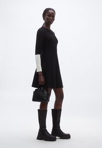 Čierne priliehavé šaty s dlhými rukávmi, biely akcent na jednom rukáve, doplnené čiernymi čižmami po kolená a malou čiernou kabelkou.
