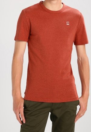 Hombre con una camiseta de manga corta lisa color óxido y pantalones verde oliva, de pie con los brazos relajados a los lados.