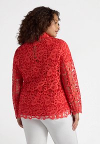 Top in pizzo rosso con maniche lunghe, caratterizzato da intricati motivi floreali e orlo scallop. Indossato sopra pantaloni bianchi, che mettono in mostra una texture dettagliata.
