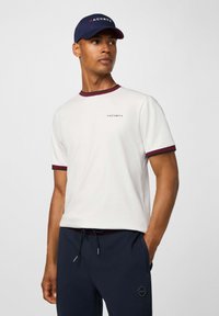 Biela tričko s bordovým lemovaním, na ľavej hrudi s logom "HACKETT". Vzájomne skombinované s tmavomodrými joggermi a zodpovedajúcou čiapkou.