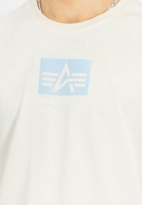 Hvid bomulds t-shirt med et lysblåt kvadratisk grafisk design, der har teksten "ALPHA INDUSTRIES" og skrå linjer, hvilket skaber et moderne udseende.