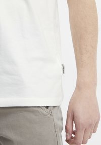 White cotton t-shirt with a side hem label, featuring short sleeves and a relaxed fit, paired with light gray pants, highlighting texture.