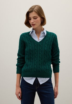 Grüner Kabelstrickpullover mit V-Ausschnitt und gerippten Bündchen, getragen über einem hellblauen gestreiften Hemd, kombiniert mit dunklen Jeans.