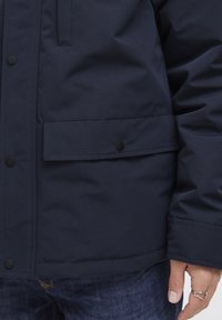 Jachetă puffer bleumarin cu o textură netedă, având o buzunar frontal cu capse și detalii de cusătură vizibile. Purtați cu blugi din denim.