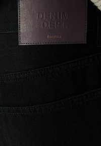 Pantalones de mezclilla negros con una etiqueta de cuero grabada con "DENIM THE DEPT." y "Bershka", destacando los detalles de artesanía y marca.