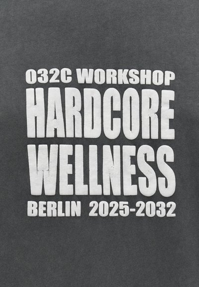 Biely tučný text na tmavom materiáli znie "032C Workshop Hardcore Wellness Berlín 2025-2032" s mierne popraskanou textúrou.