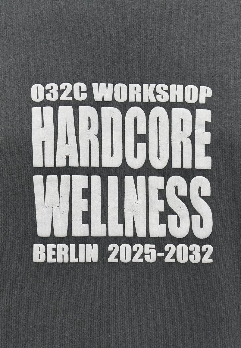 Weiße, fettgedruckte Schrift auf dunklem Stoff mit der Aufschrift "032C Workshop Hardcore Wellness Berlin 2025-2032" und einer leicht rissigen Textur.