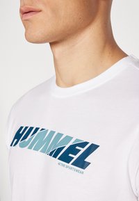 Uomo che indossa una maglietta bianca con una grande scritta blu "HUMMEL" e la dicitura "ACTIVE SPORTSWEAR" stampata sotto sul petto, collo e linea della mascella visibili.