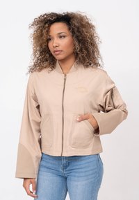 Veste beige courte avec fermeture éclair à l'avant, deux poches avant et manches larges. Tissu lisse avec des coutures ton sur ton et un détail de logo brodé.