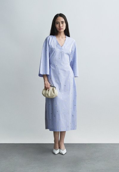 Φόρεμα μακρύ, ανοιχτό μπλε με ρίγες, V-λαιμόκοψη και μανίκια τριών τετάρτων, με χρυσή κεντητική. Συνδυασμένο με κρεμ τσάντα με πιέτες.