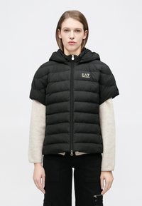 Fekete rövid ujjú steppelt dzseki kapucnival, cipzáros elülső résszel és arany logóval a mellkasnál. Az anyaga sima és könnyűnek tűnik.