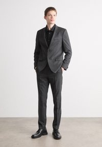 Heren donkergrijs maatpak met een getailleerde fit, gemaakt van een gestructureerde stof. Inclusief een enkelgeknoopt jasje en bijpassende broek, aangevuld met zwarte schoenen.