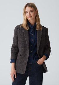 Blazer a quadri marrone in tessuto testurizzato, con vestibilità sartoriale, revers a scotto e due tasche frontali, indossato sopra una camicia in denim blu navy.