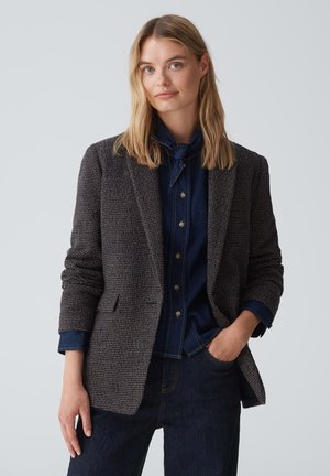 Blazer à carreaux marron en tissu texturé, avec une coupe ajustée, des revers en pointe et deux poches avant, porté sur une chemise en denim bleu marine.