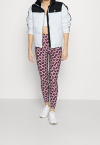 Kurtka puchowa o białym korpusie i czarnych rękawach, w połączeniu z wzorzystymi legginsami w kolorze burgundowym i białym oraz białymi butami sportowymi.