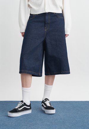 Isik, kes kannab tumedat teksasid laiade säärega lühkareid, valgeid sokke, mustvalgeid tossusid ja valget nööbitavat kampsunit.