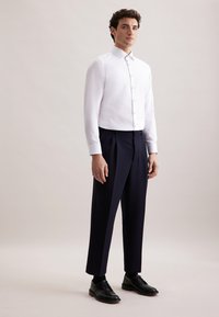 Uomo in camicia bianca con bottoni, pantaloni neri su misura, scarpe eleganti nere, contro uno sfondo neutro semplice.