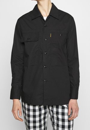 Person, die ein schwarzes, langärmeliges Hemd mit Brusttaschen und schwarz-weiß karierte Hose trägt.